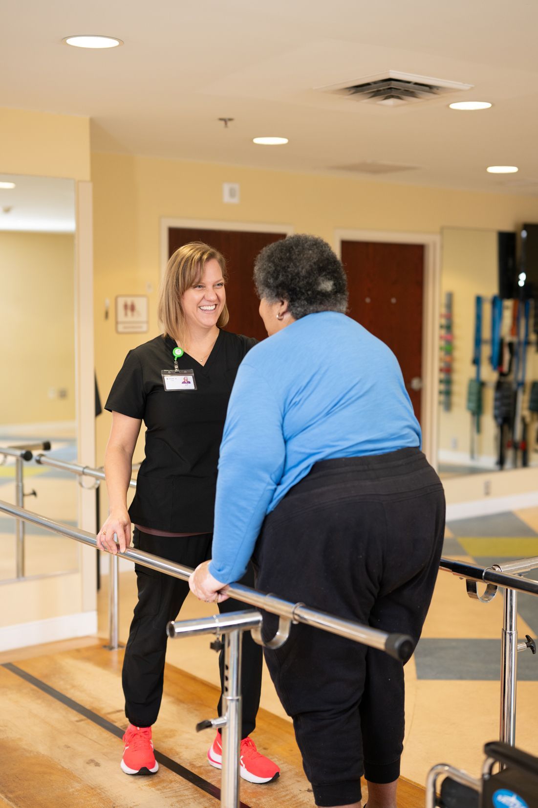 A woman is helping a woman in a wheelchair walk on parallel bars.