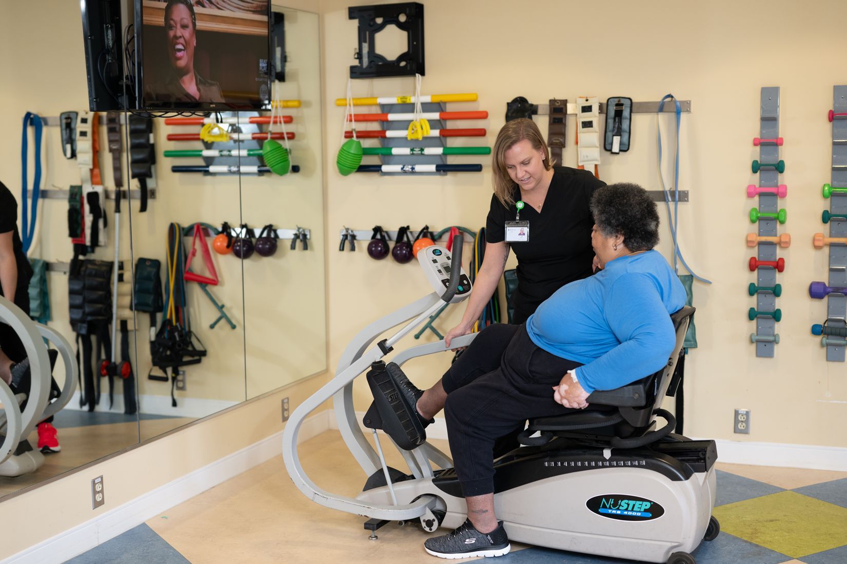A woman is helping a woman on an exercise bike in a gym.