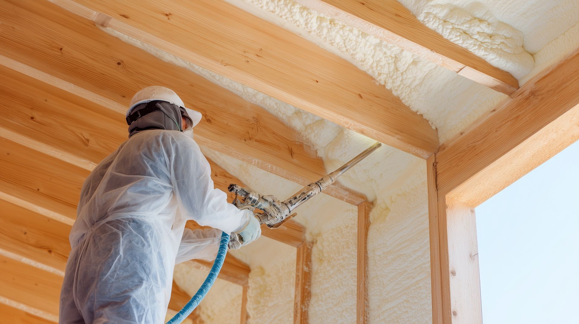 A worker in a white protective suit uses a spray gun to apply insulation foam between wooden ceiling beams.