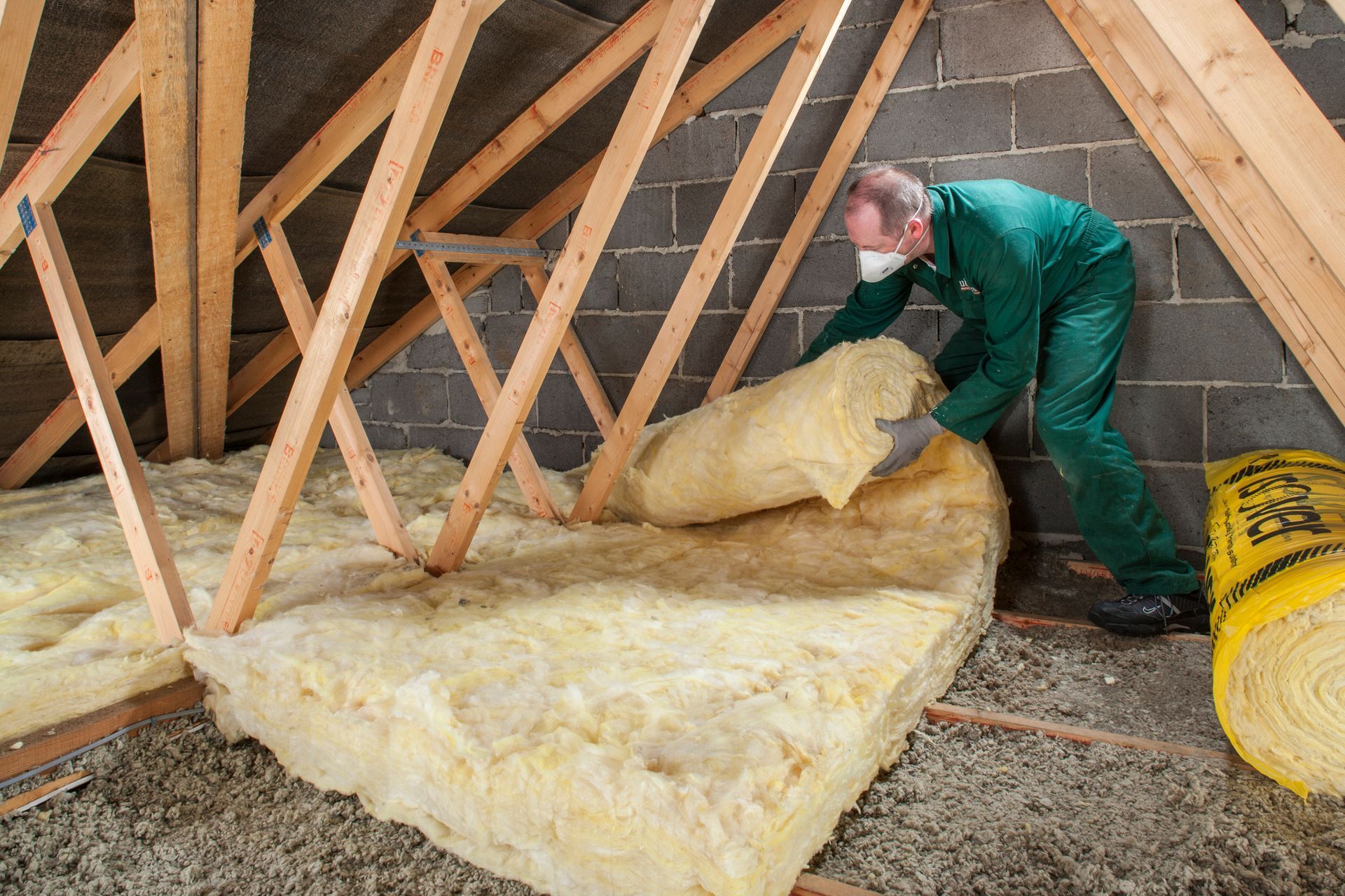 A man in a green bodysuit lays a layer of insulation material in an attic.