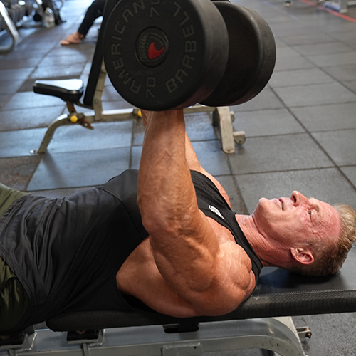 Ron Matthews is laying on a bench lifting a dumbbell that says american body
