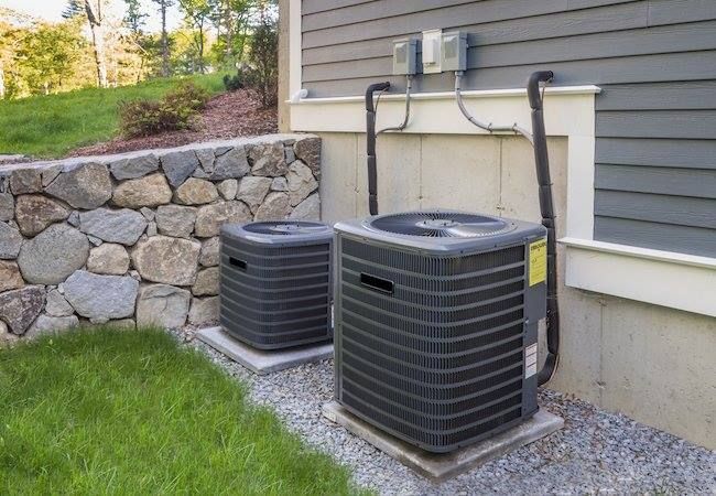 Two air conditioners are sitting outside of a house next to a stone wall.