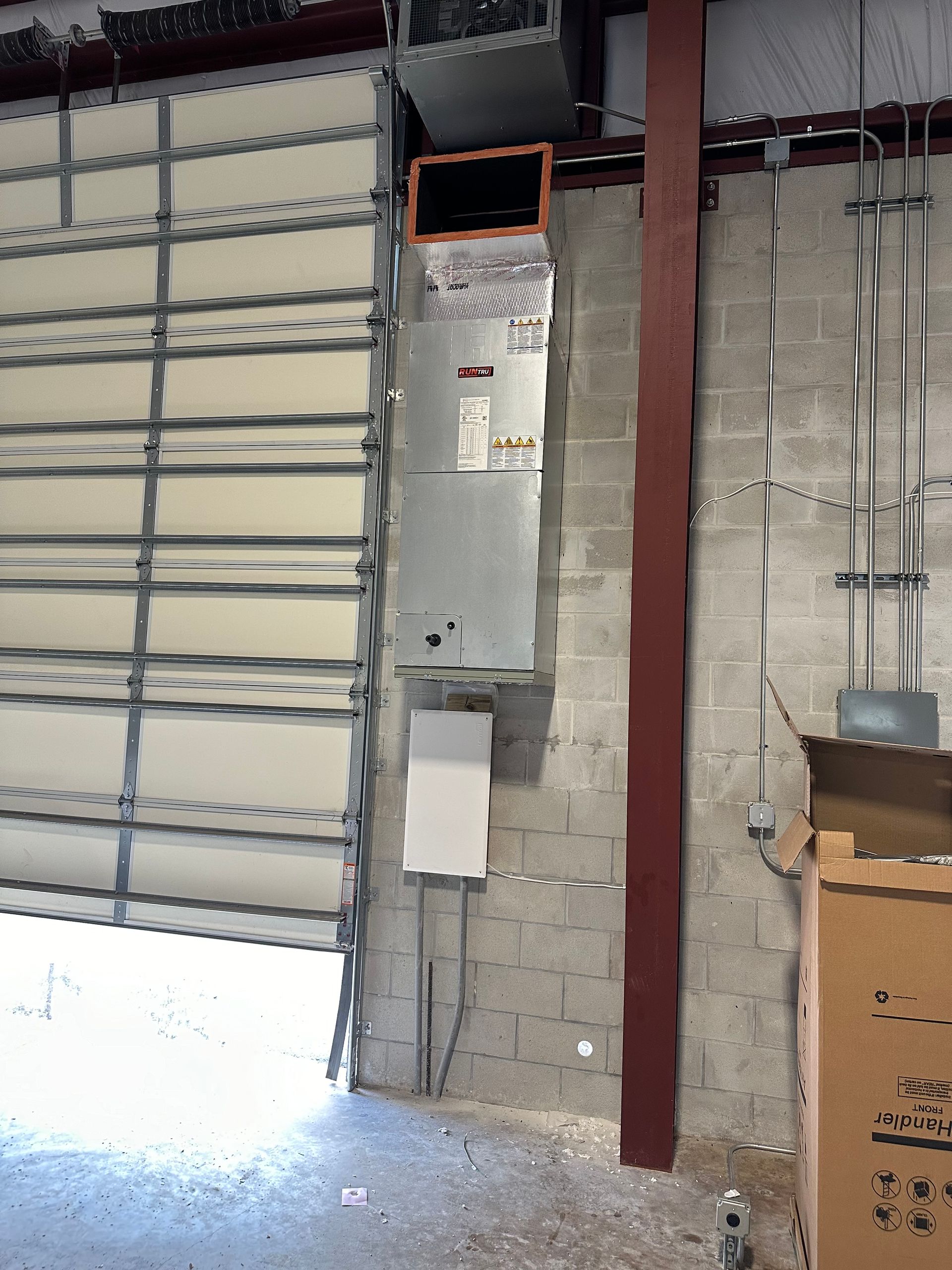 Large garage door open beside an air conditioning unit, mounted on a concrete block wall.