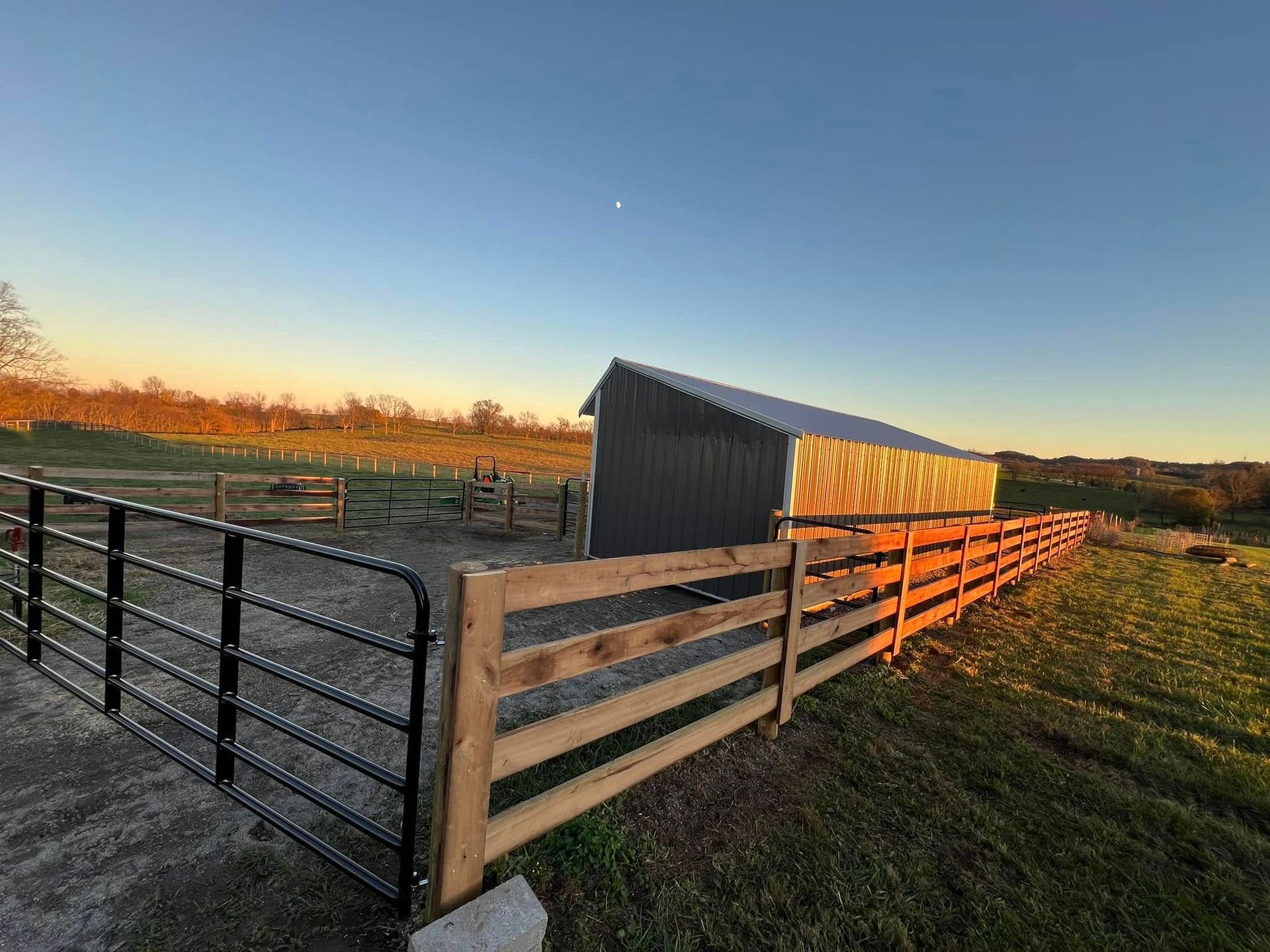 A small gray barn with a metal roof sits in a fenced pasture at sunset, casting long shadows.