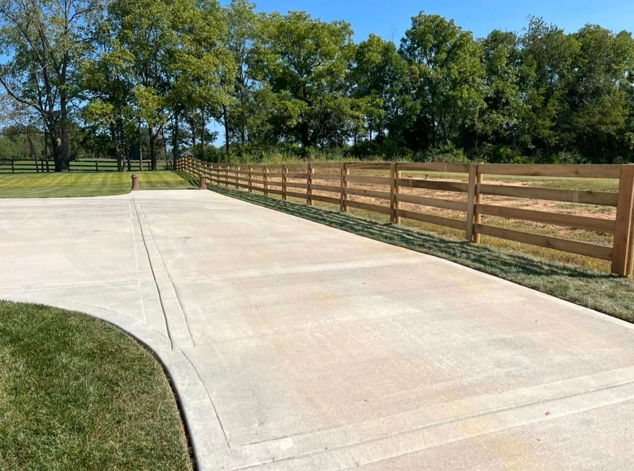 Concrete driveway with wooden fence alongside green lawn and trees under a blue sky.