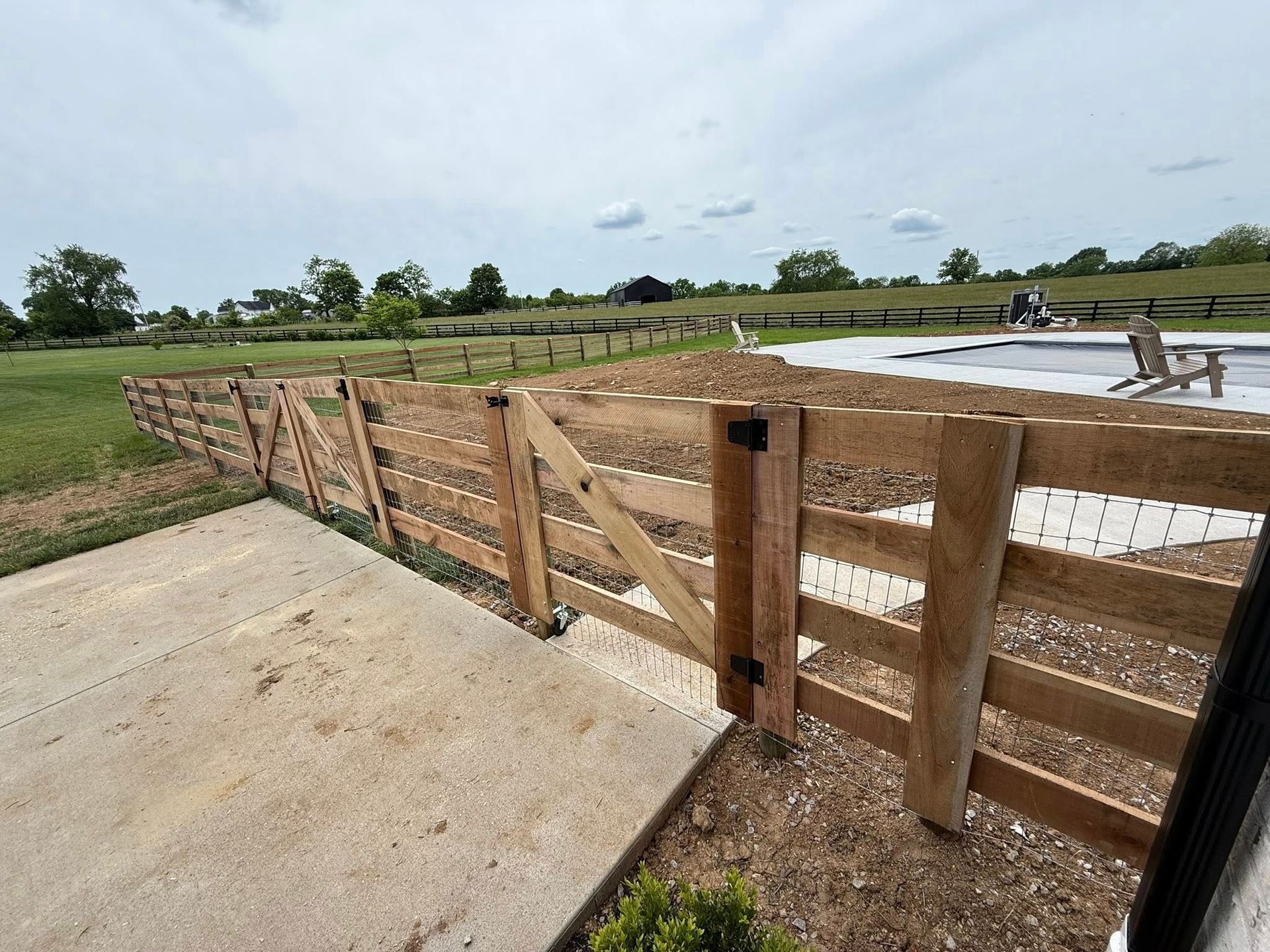 Wooden fence with gates, separating a concrete patio from a grassy yard, with a swimming pool visible in the background.
