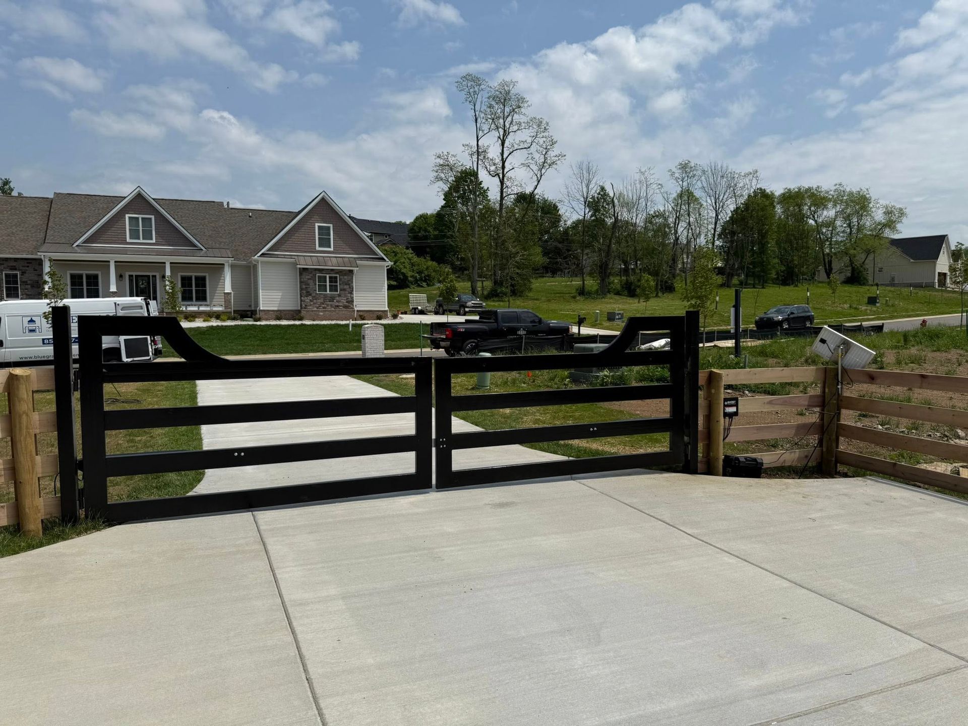 Black wooden gates open onto a concrete driveway, leading to a neighborhood with houses and parked cars.