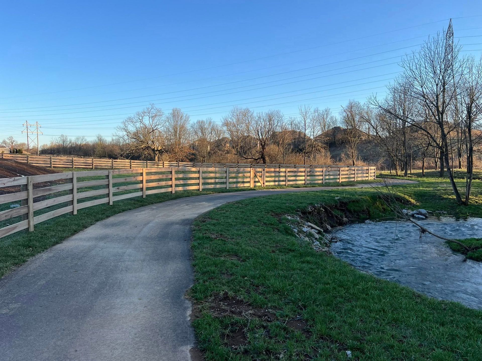Paved pathway curving along a grassy bank with a wooden fence, creek, and trees under a blue sky.