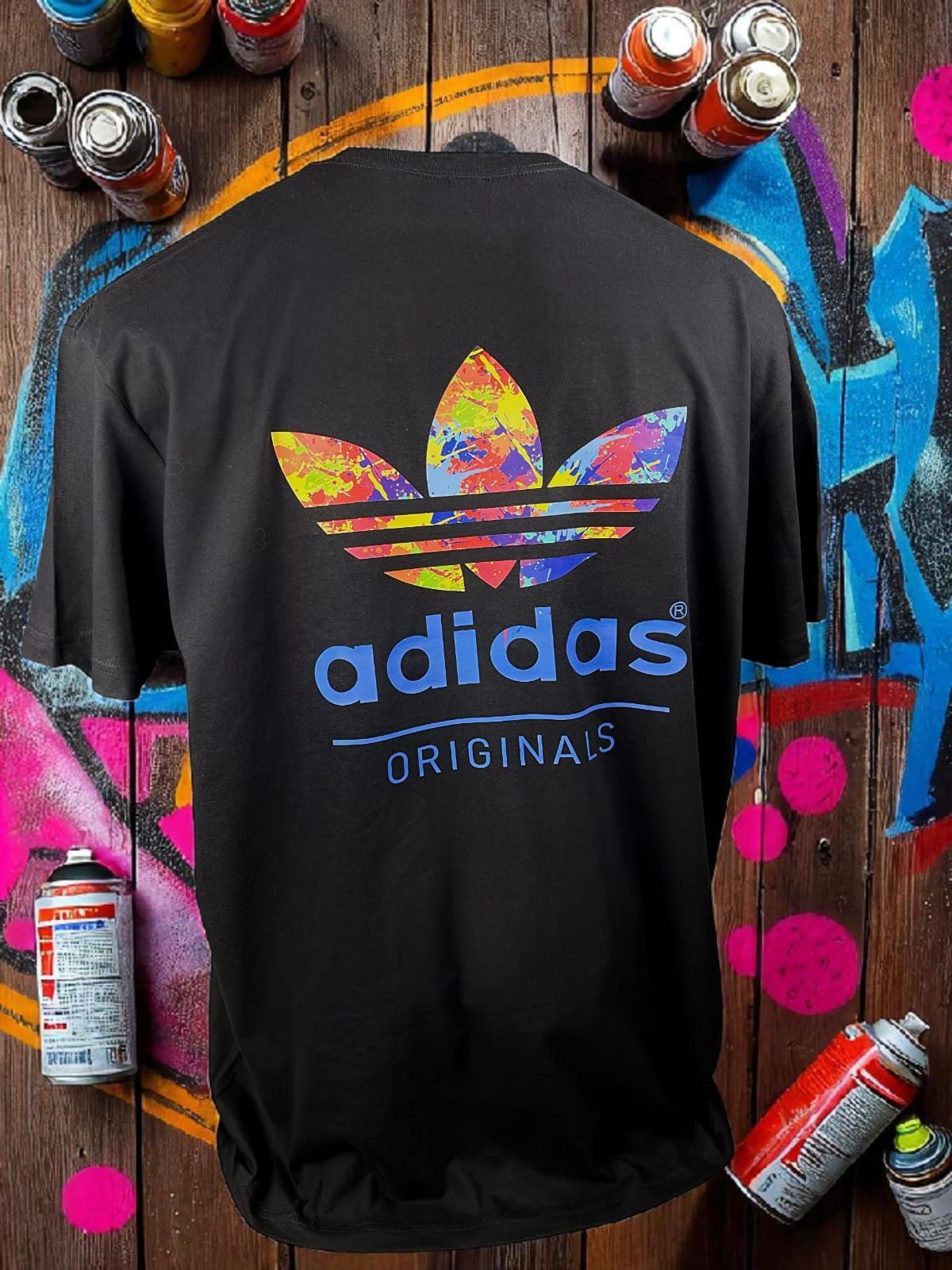 Camiseta negra de Adidas Originals con un colorido logotipo de trébol y texto azul sobre un fondo de graffiti.