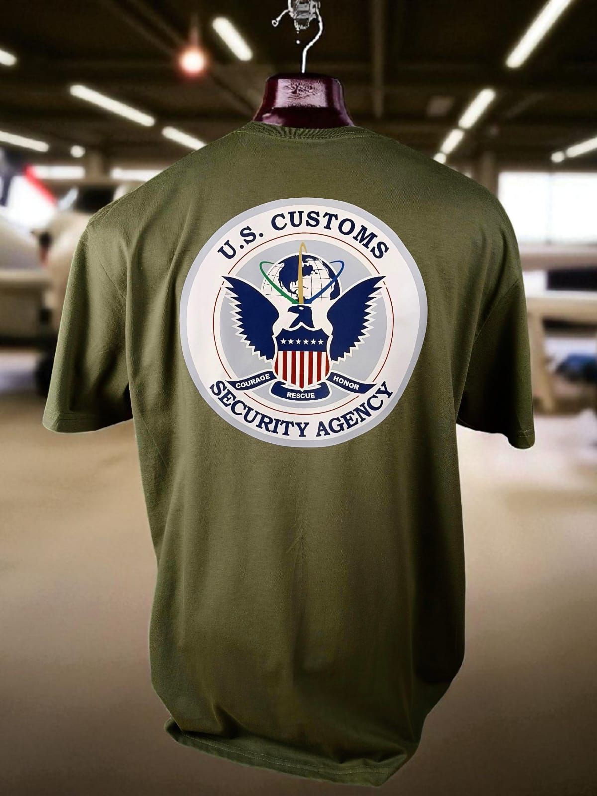 Camiseta verde oliva de la Agencia de Seguridad Aduanera de EE. UU., expuesta en una percha. El emblema está en la espalda.