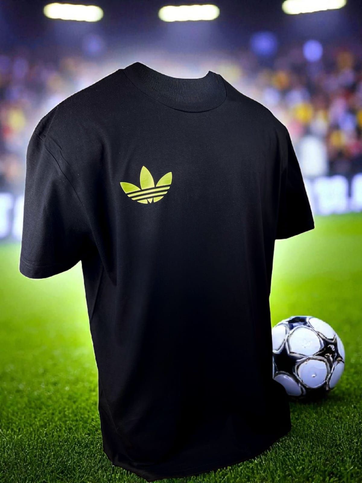 Camiseta Adidas negra con logo amarillo, en un campo de fútbol, ​​con un balón de fútbol.