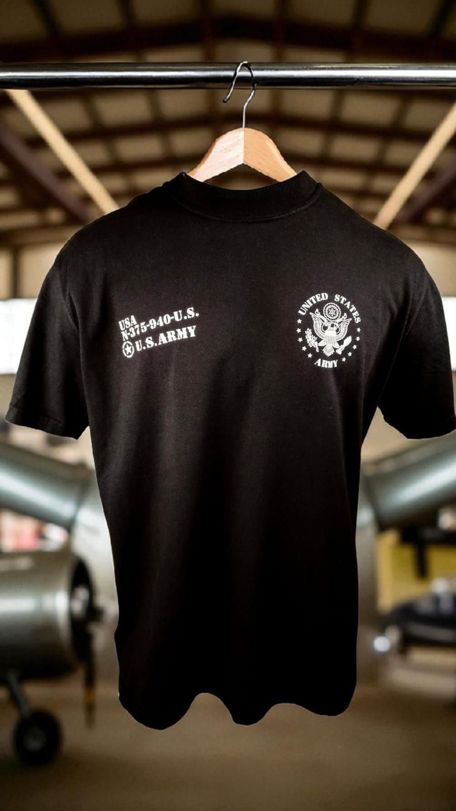 Camiseta negra con el logotipo y el texto del ejército de EE. UU., colgada en un perchero en un hangar de aviones.