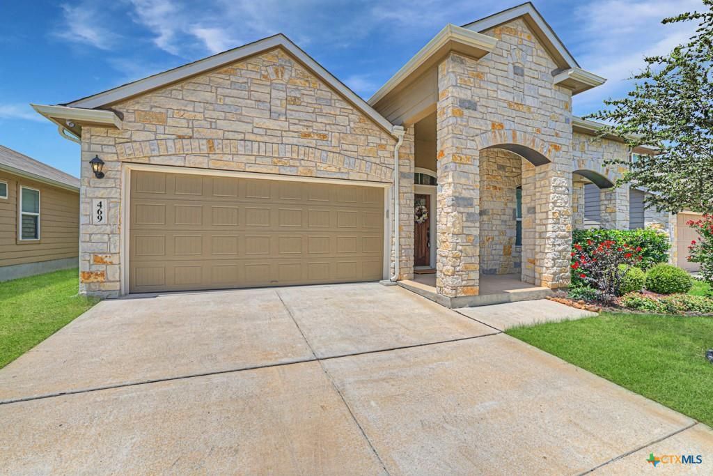 Stone-clad house with attached garage and arched entry. Concrete driveway and green lawn under a blue sky.