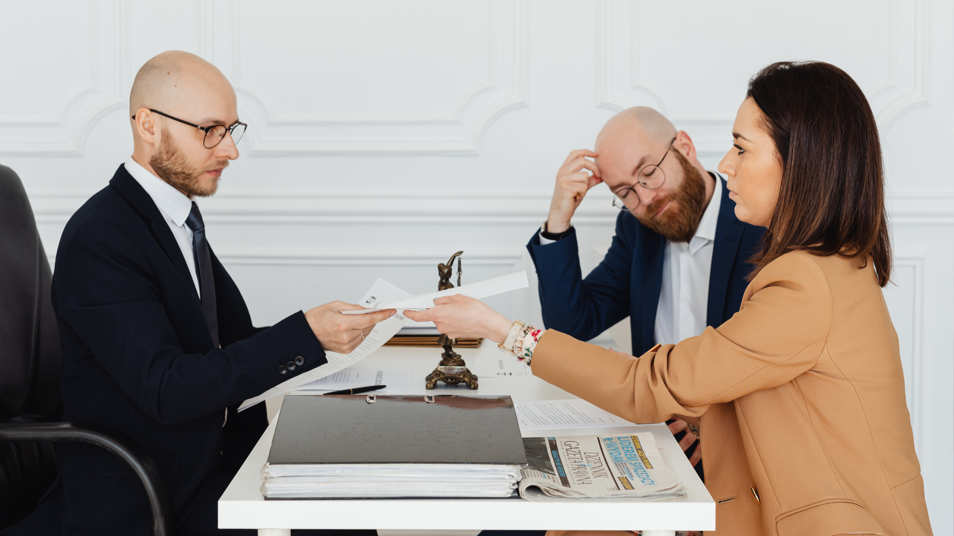 Three people in a meeting discussing documents across a desk, with one person handing over papers.