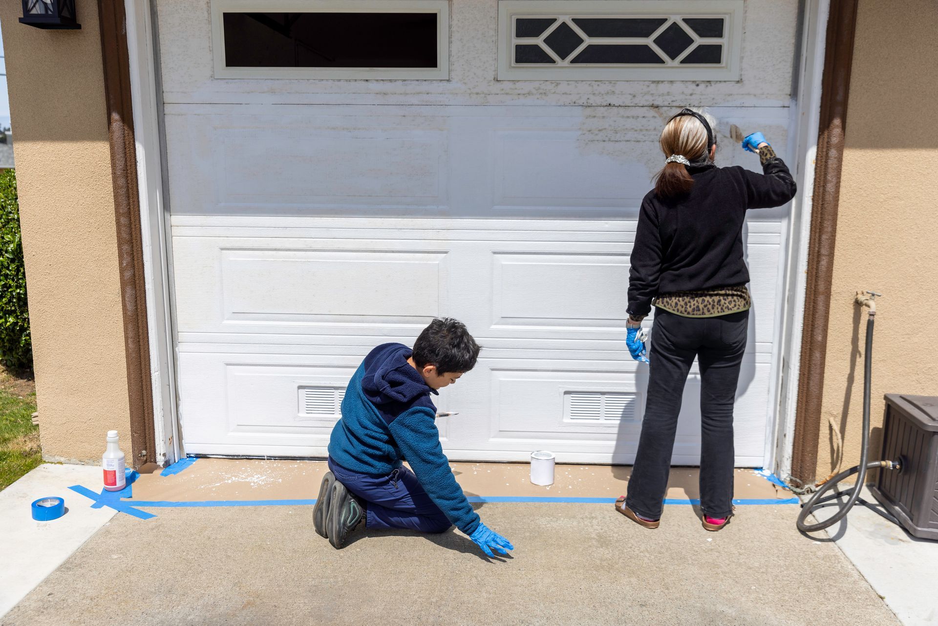 A pair of garage door company workers paint a garage door white in front of house curb.