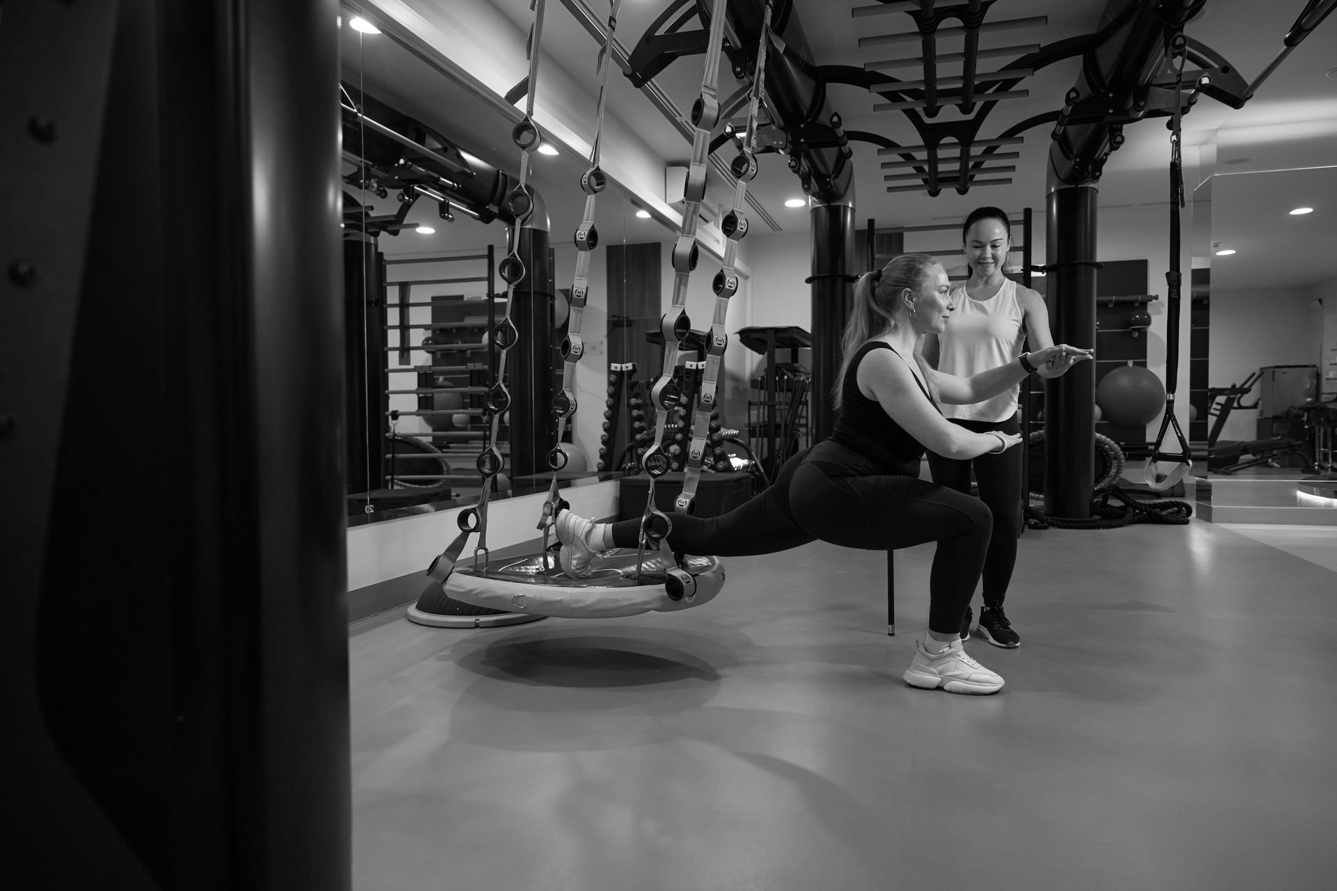 A man is helping a woman do squats in a gym.