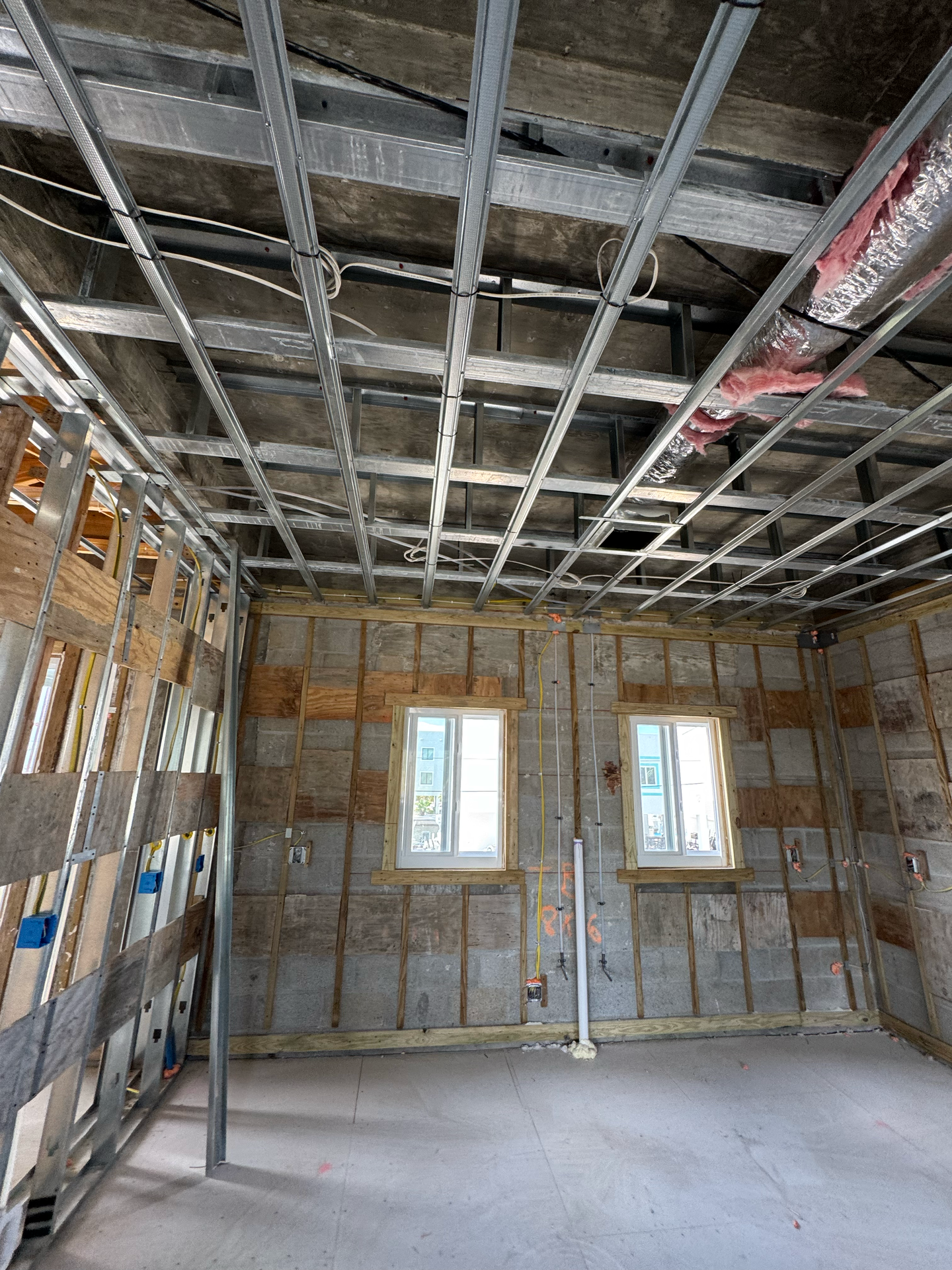 Interior of a room under construction with metal framing for walls and ceiling. Two windows and concrete block walls are visible.