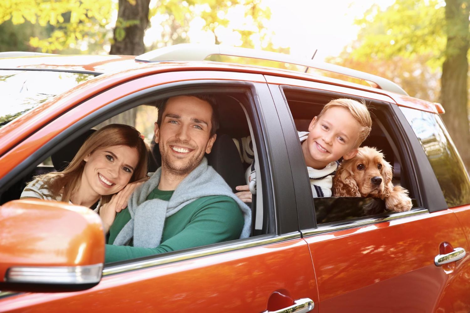 Smiling family with dog leaning out of an orange car window in bright sunlight
