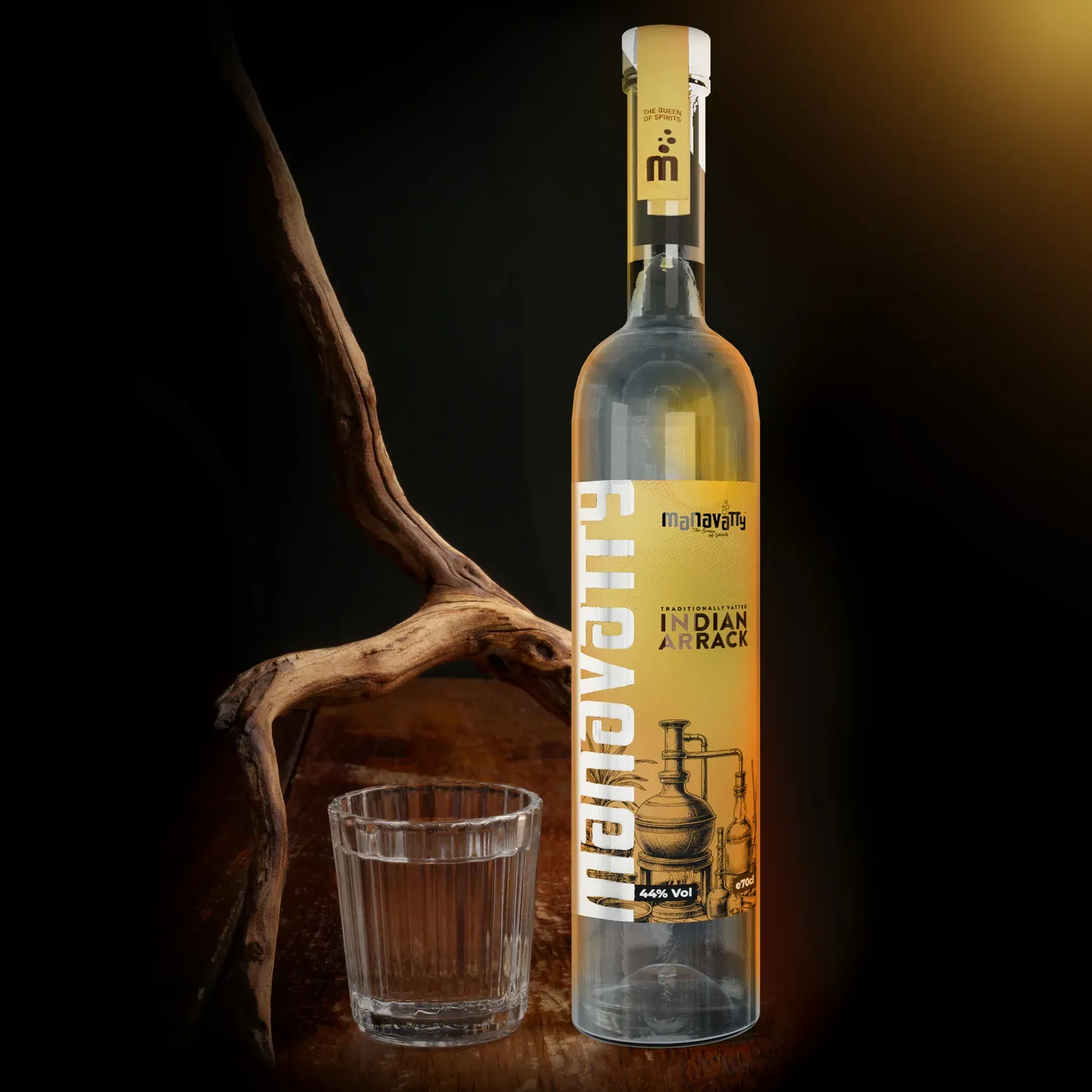 A tall bottle of Manavanth Indian Arrack next to a glass of clear liquid on a wooden table with a branch in the background.