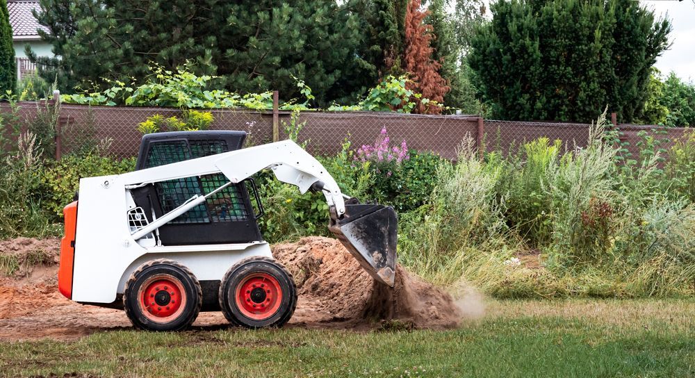 White Bobcat skid-steer loader dumping soil onto grass near a fence and bushes.