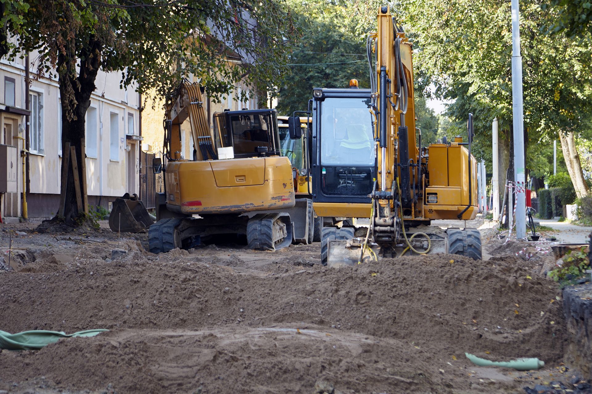 Two yellow construction excavators on a dirt road next to a building and trees.