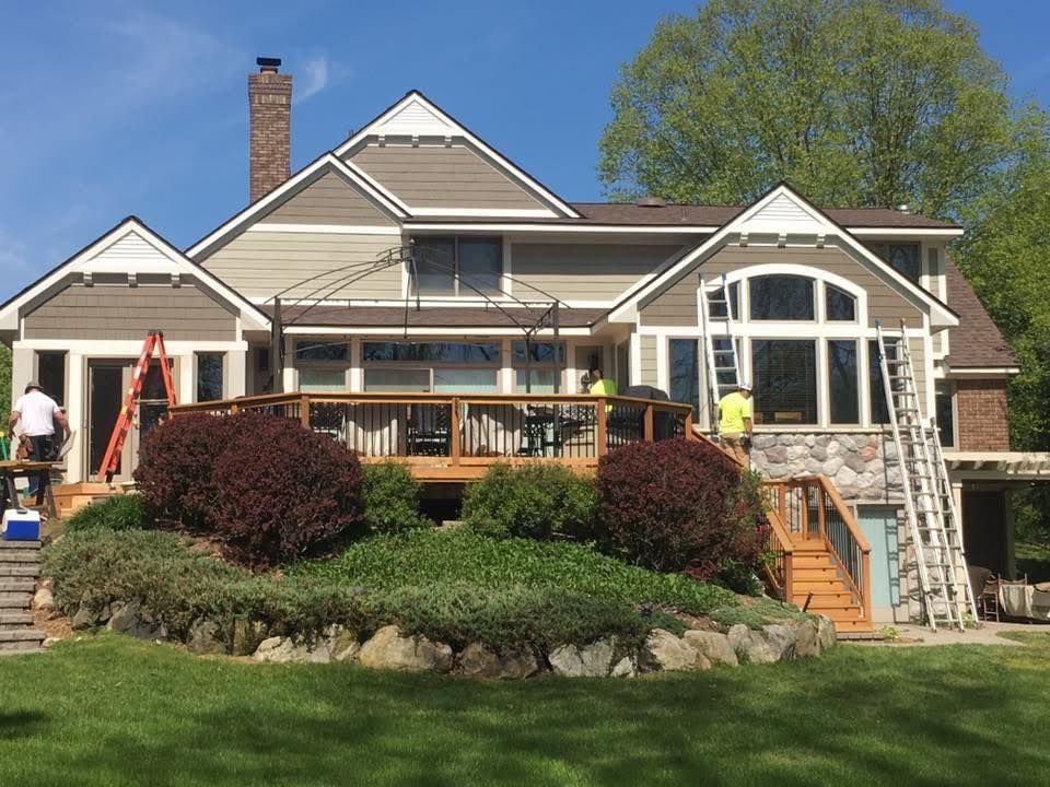 Back of a house being painted; deck, landscaping, two workers on ladders, green lawn, blue sky.
