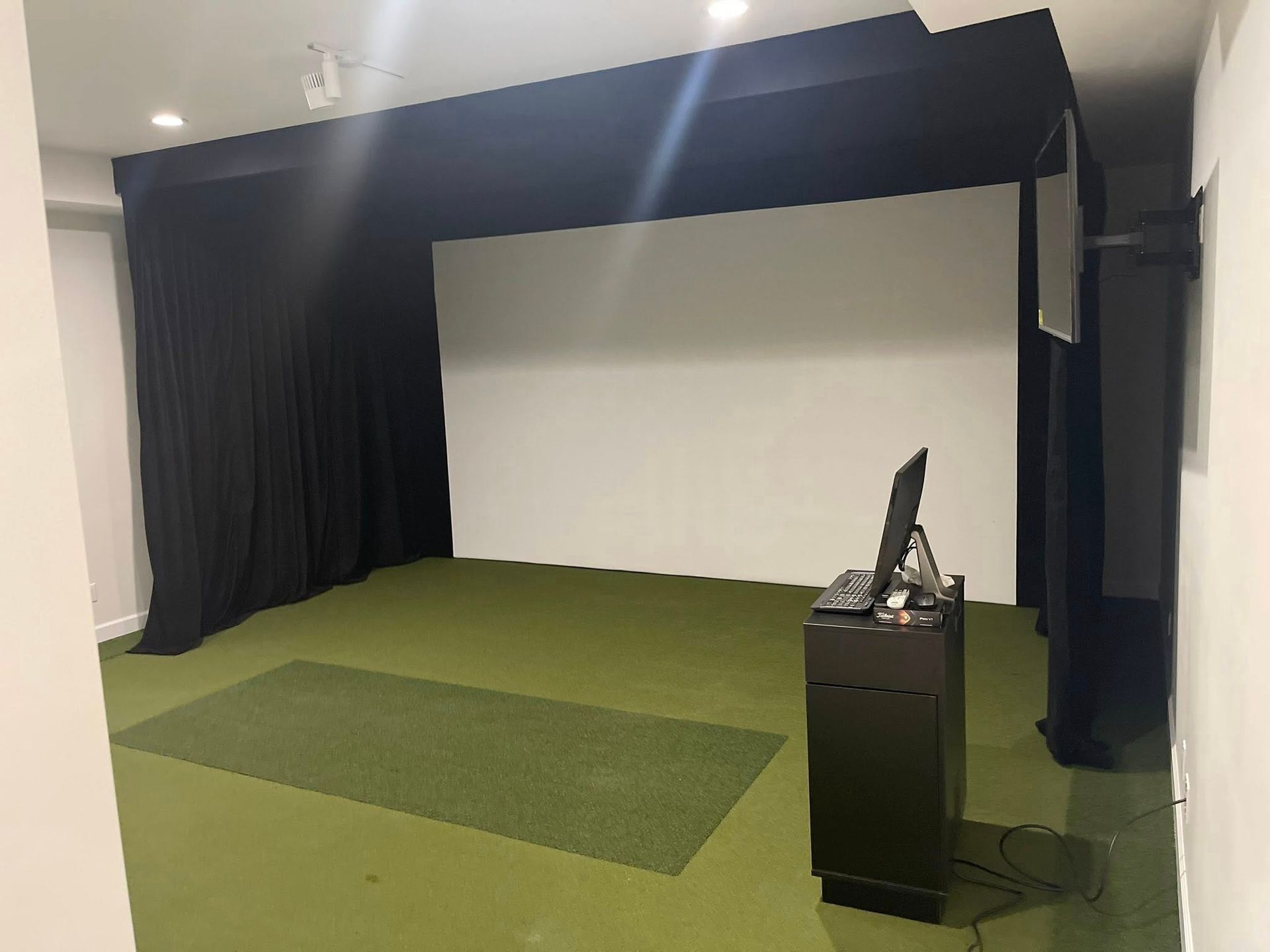 Indoor golf simulator setup with green turf, white screen, and black curtains. A computer monitor sits on a black stand.