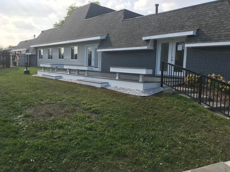 Gray building with multiple entryways, a deck with benches, and a grassy yard.