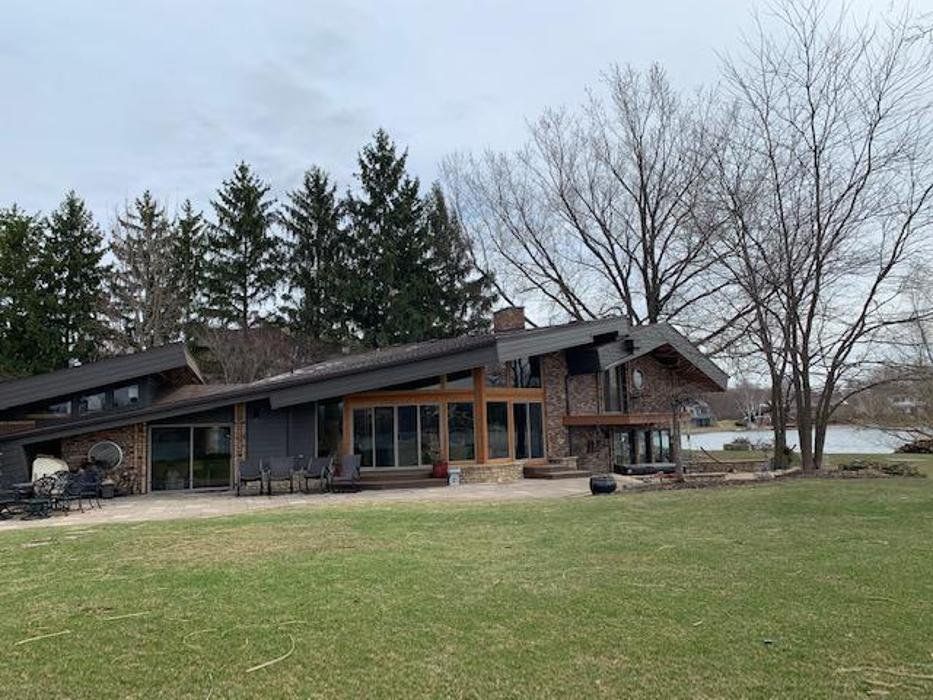 Modern, low-slung house with angled roof, large windows, and stone facade, set on grassy lawn near water and trees.