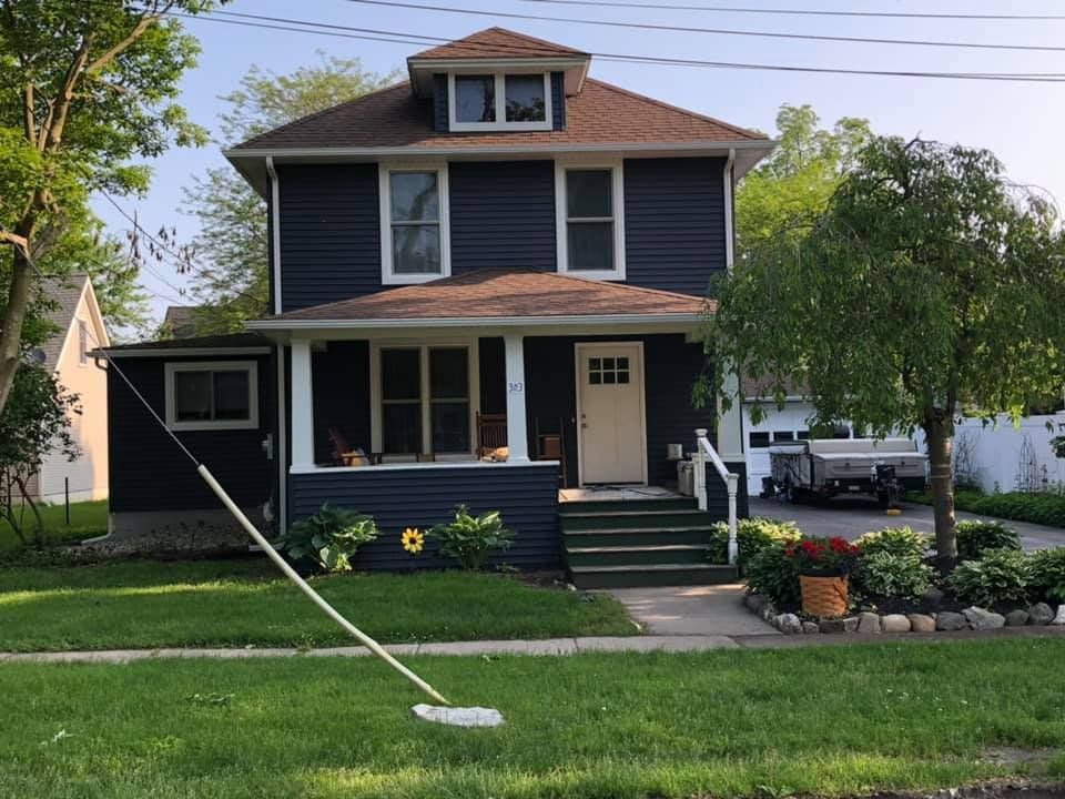 Two-story blue house with porch, green lawn, and small garden. Brown roof, white trim, and a tree on the right.