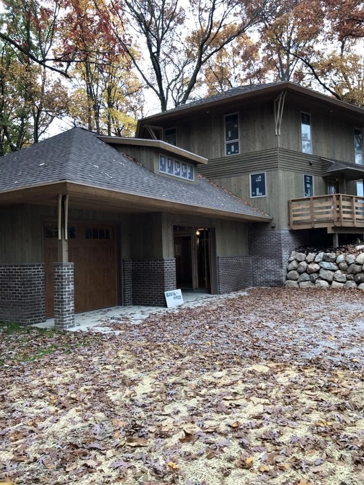 Two-story house with attached garage, brown siding, wooden deck, stone base, autumn leaves on the ground.