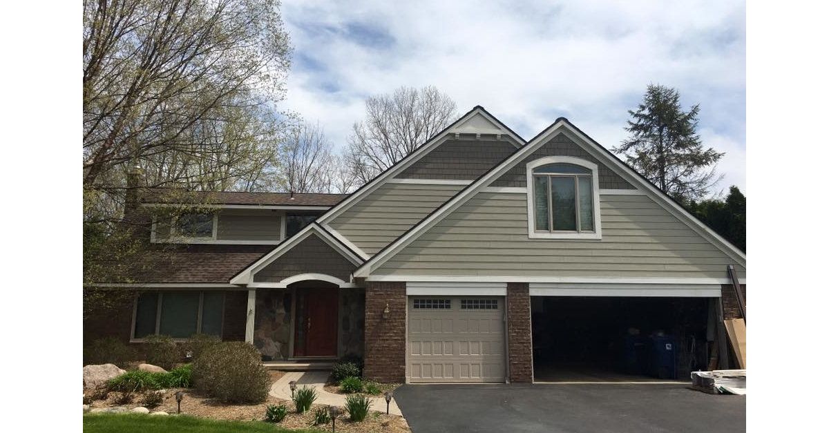 House with brown roof, green siding, brick accents, and a two-car garage. Overcast sky.