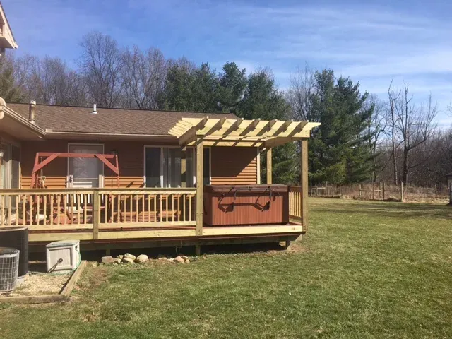 Wooden deck with hot tub under a pergola, next to a house with a grassy yard and trees.