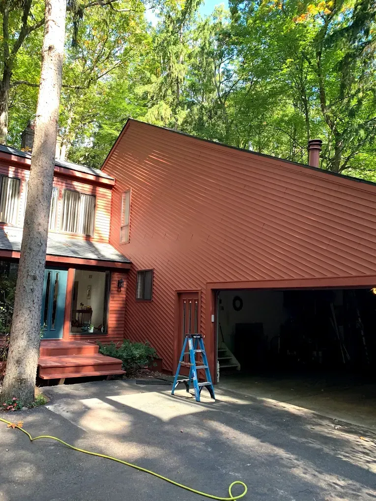 Red-sided house with attached garage, blue ladder, and a black asphalt driveway, surrounded by green trees.