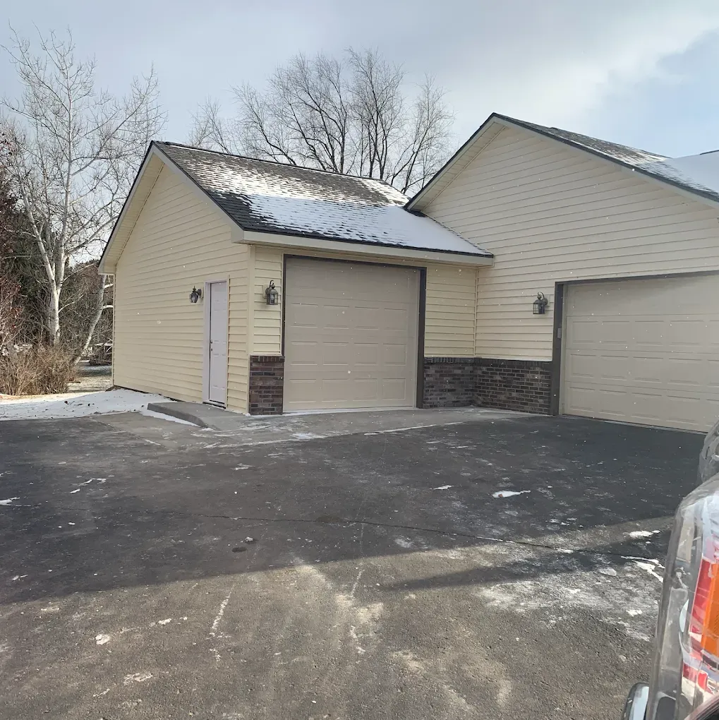 Light yellow detached garage with beige doors. Stone veneer accents. Pavement, snow, and overcast sky.