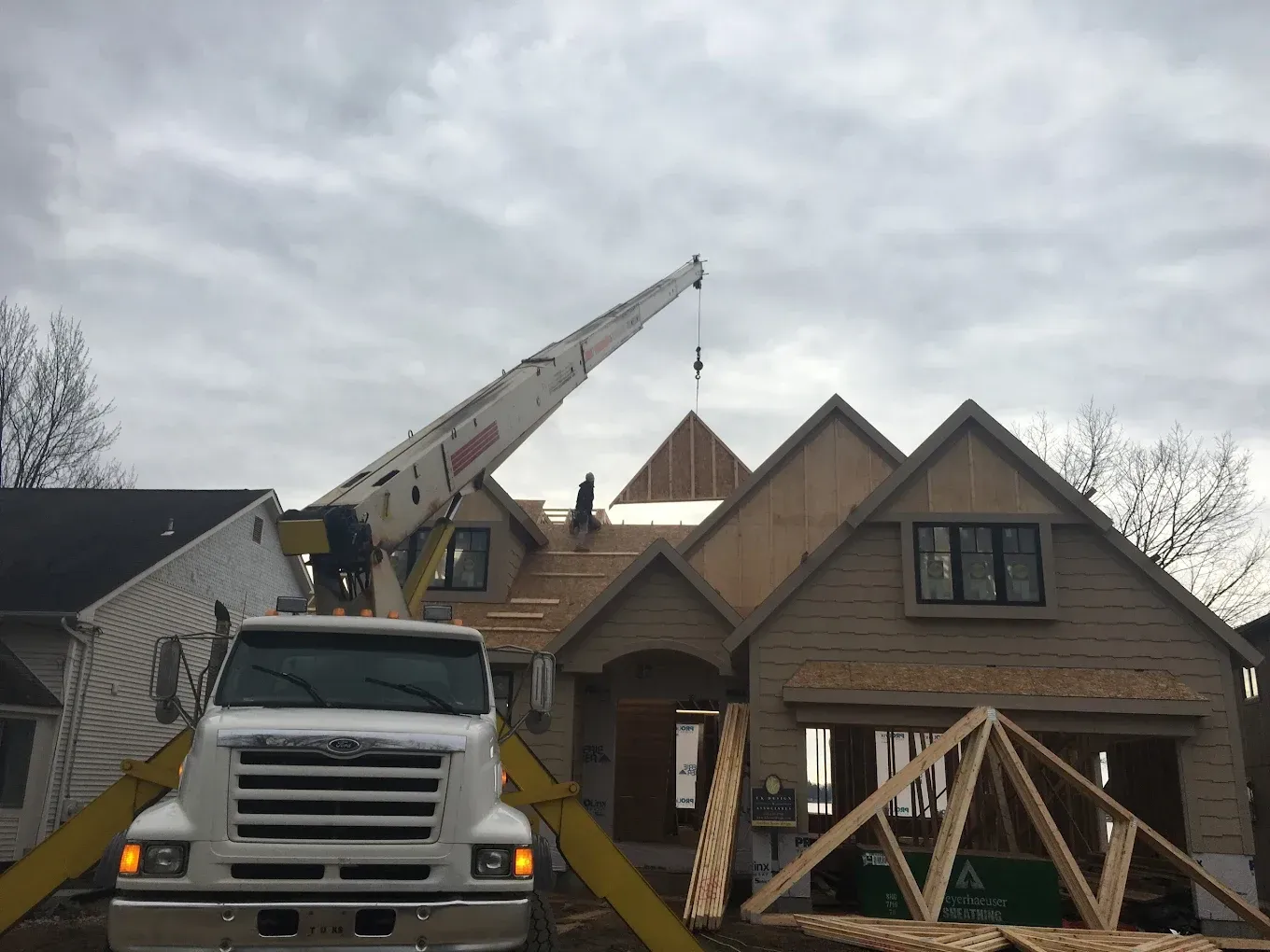 Construction site. A crane lifts roof trusses onto a partially built house with exposed wood framing.