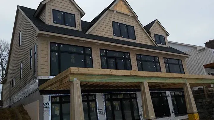 Three-story house under construction with light brown siding, black windows, and a wooden porch.