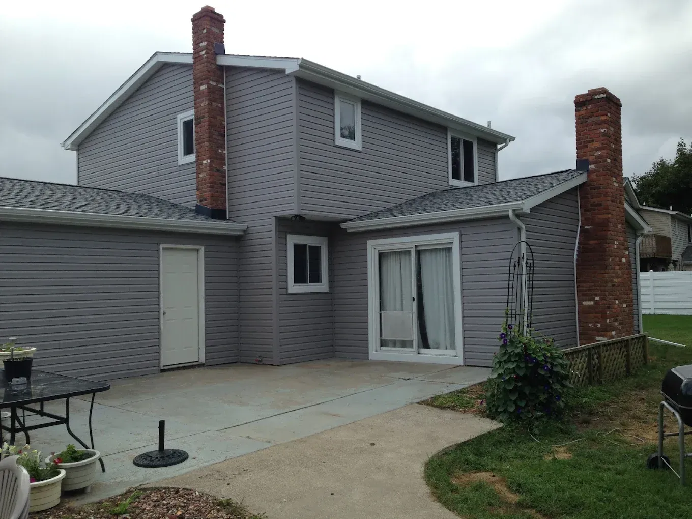 Back of a two-story gray house with brick chimneys, patio, white door, and sliding glass door. Overcast sky.