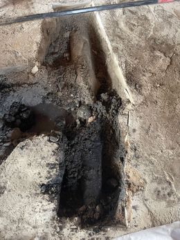 An excavation pit revealing dark soil and objects in a sandy environment, potentially an archeological site.