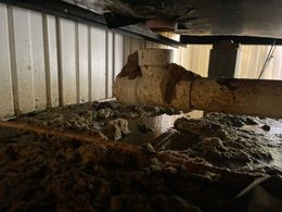 Overflowing sewage pipe under a structure with sludge on the ground and white paneling.