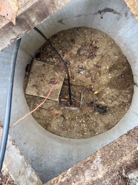 A concrete access box with a circular lid covered in sand and wires.