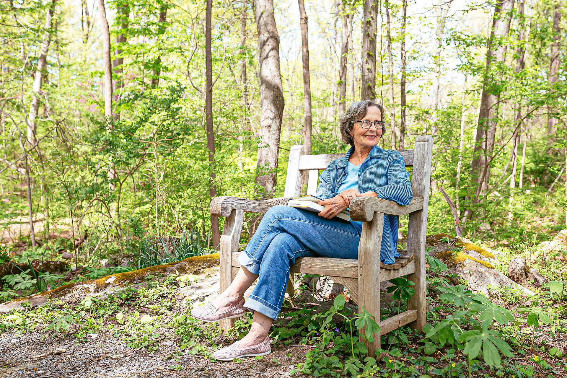 A woman is sitting on a wooden bench in the woods reading a book.