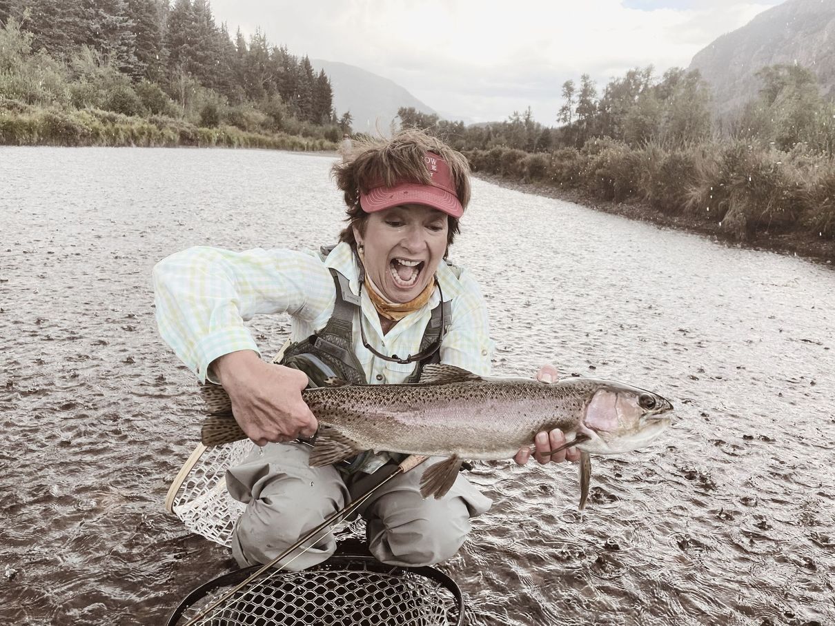Woman with a very excited look n her face and a red visor in the middle of a river with a HUGE fish she just caught on a Zia Fly Guided Fishing Trip.