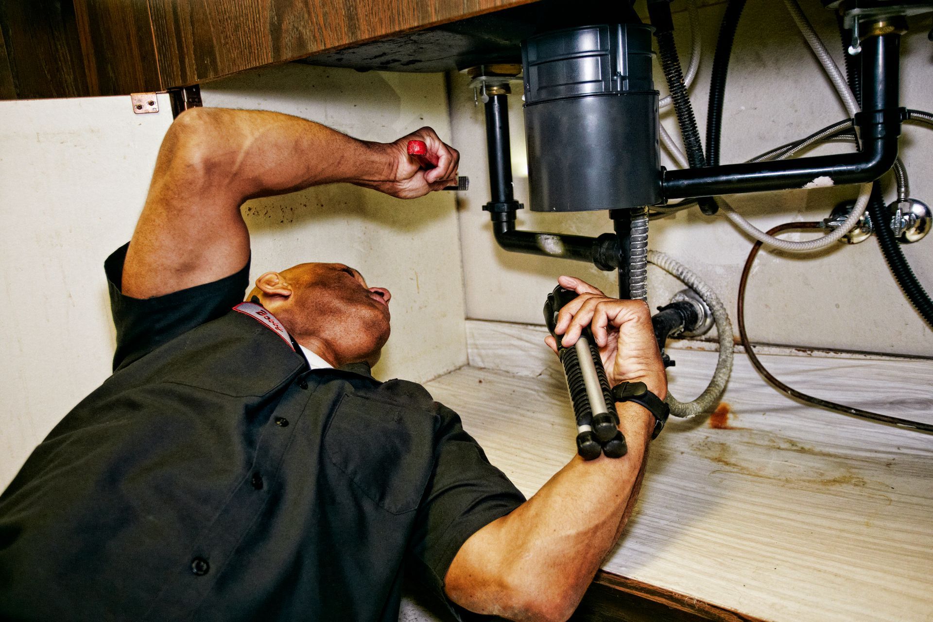 Plumber working under a kitchen sink, using a flashlight and wrench.