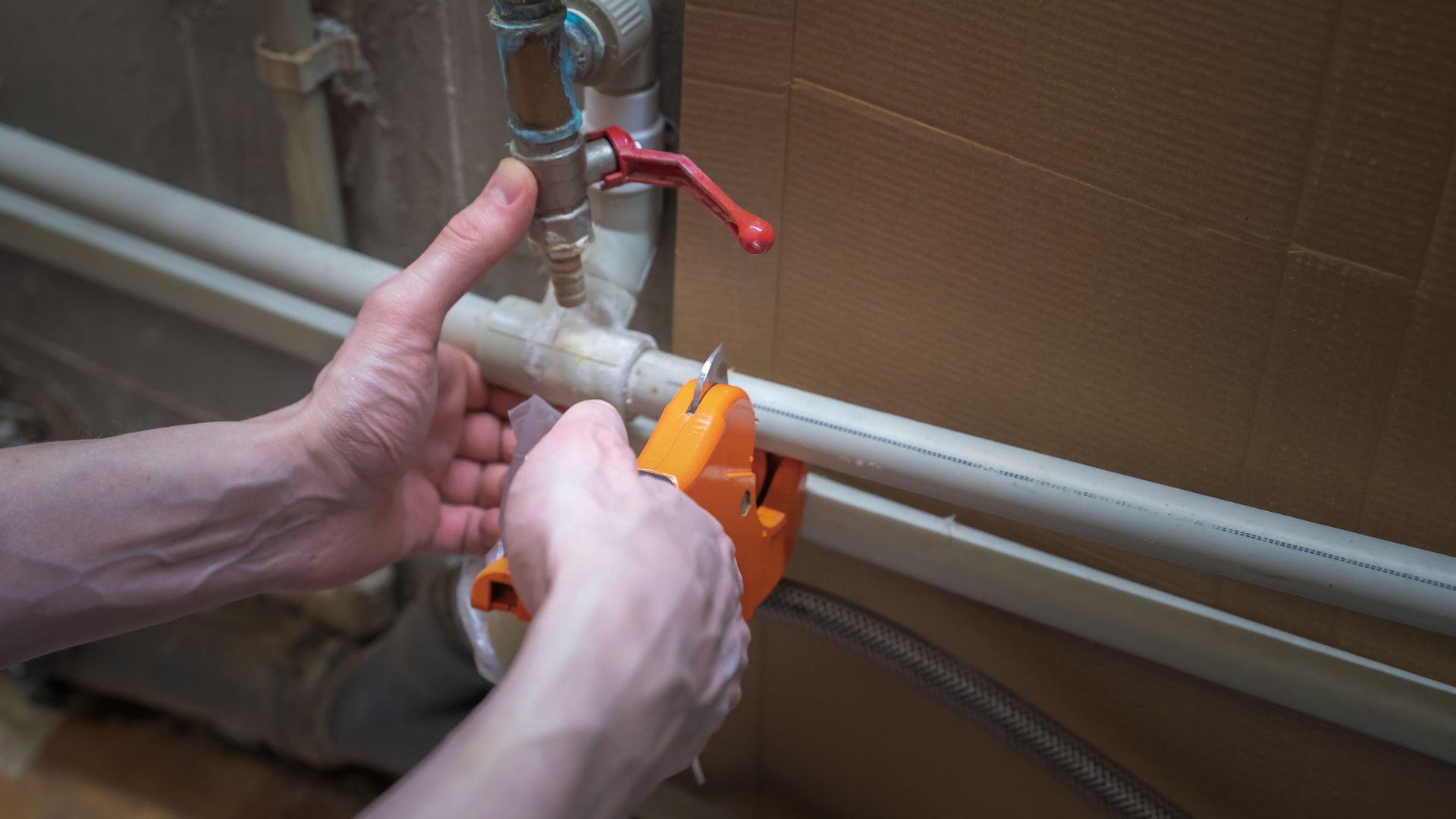 Person using an orange pipe cutter to cut a white plastic pipe near a red-handled valve.