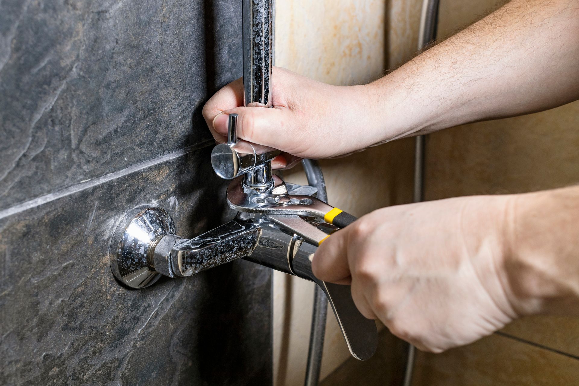 Hands using a wrench to repair a chrome shower fixture on a tiled wall.