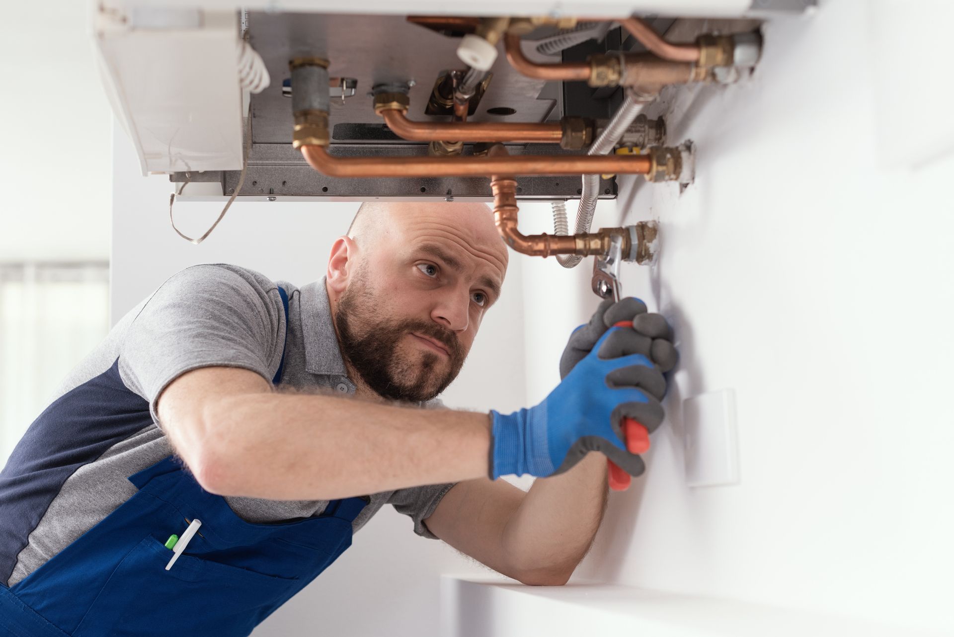 Plumber in blue gloves, repairing a boiler with a wrench. White wall, copper pipes visible.