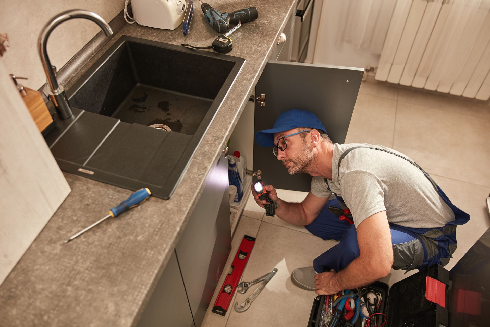 Plumber inspecting sink plumbing with a flashlight. He's wearing a blue cap and overalls, looking inside a cabinet.
