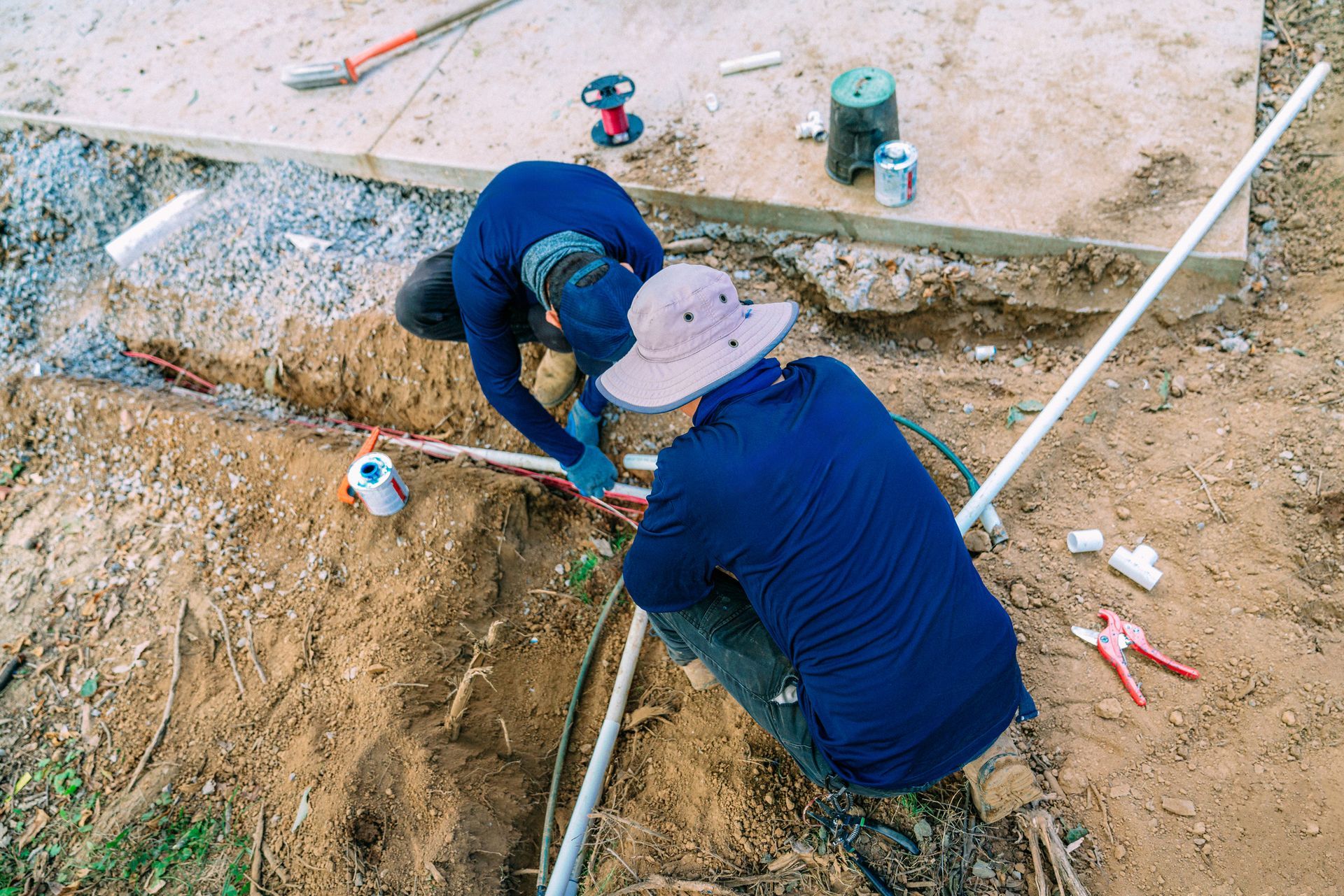 Two people in blue shirts working on underground pipes in a dirt trench, near a concrete slab.