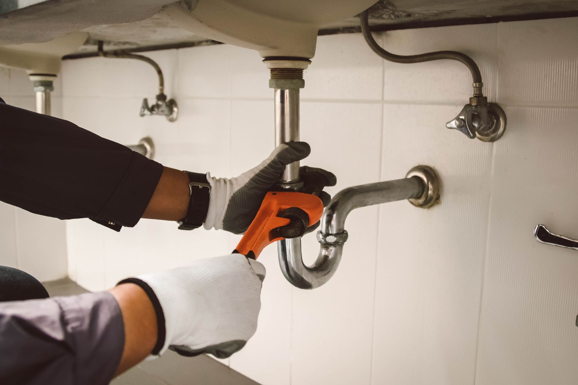 Plumber with wrench working on a bathroom sink's drain pipe, hands in gloves, white tile background.